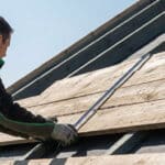 Un couvreur mesure avec un mètre ruban des planches de bois installées sur la charpente d'un toit en construction.