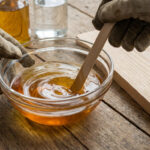 Gants de travail mélangeant un liquide ambré dans un bol en verre sur une table en bois avec une planche et un pinceau.
