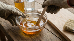 Gants de travail mélangeant un liquide ambré dans un bol en verre sur une table en bois avec une planche et un pinceau.