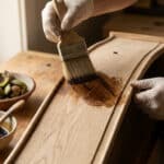 Artisan appliquant du brou de noix sur un tiroir en bois avec un pinceau, près d'un bol de noix fraîches.