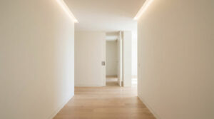 Un couloir lumineux et épuré avec des murs blancs, un sol en parquet en bois clair et un éclairage indirect au plafond.