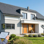 Un expert utilise un télémètre laser pour mesurer la hauteur d'une maison moderne avec un plan sur une tablette au premier plan.