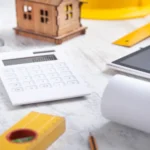 Calculatrice blanche près d'une maquette de maison en bois, d'un niveau à bulle, d'un mètre ruban et de plans roulés