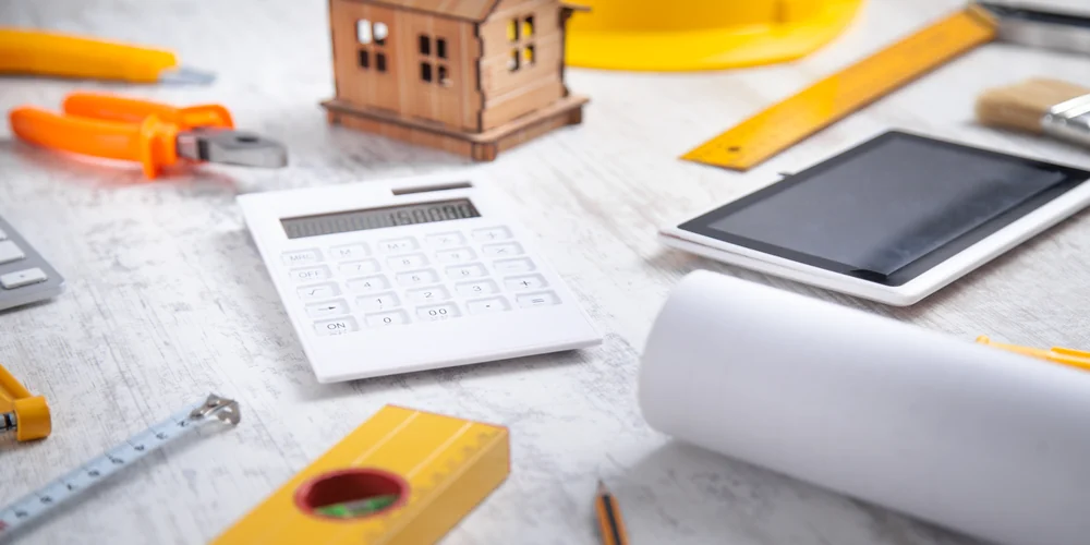 Calculatrice blanche près d'une maquette de maison en bois, d'un niveau à bulle, d'un mètre ruban et de plans roulés