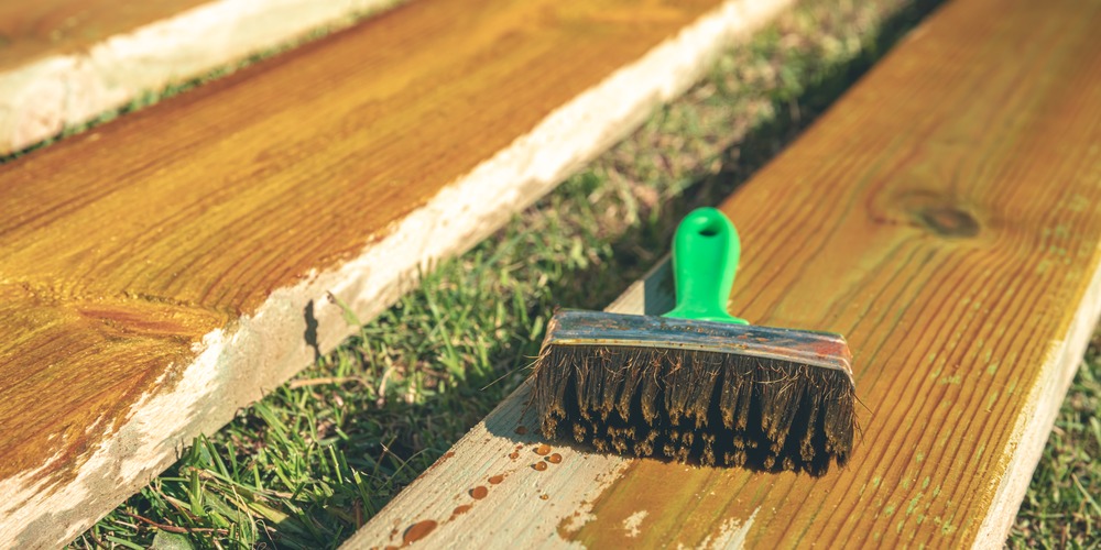 Pinceau posé sur des planches en partie enduites d'un produit de protection, dans l'herbe d'un jardin